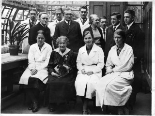 Ein Gruppenbild zum Dienstjubiläum der älteren Frau, die in Zivilkleidung vorne im Zentrum sitzt, aufgenommen 1935. Die Frauen neben ihr tragen weiße Arbeitskittel, die Männer hinter ihnen stehen und tragen einen Anzug, eine Frau im Kostüm steht ebenfalls hinter den sitzenden Frauen.