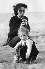 Mira Lobe, eine junge Frau mit kinnlangem schwarzen Haar, sitzt hockend am Strand mit einer Blockflöte im Mund. Vor ihr sitzt spielend ein kleines Mädchen.