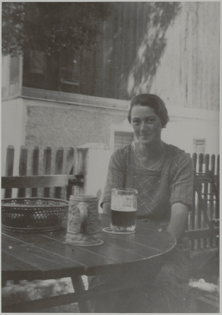 Das Bild zeigt eine Frau sitzend in einem Gastgarten mit einem Glas Bier.